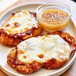 Golden breaded chicken breast topped with melted Swiss cheese and smoked ham on a white plate beside mustard sauce