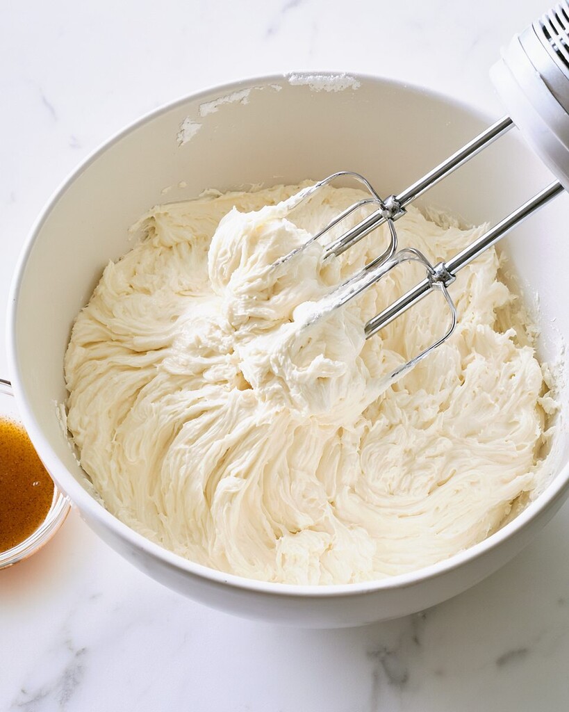 Thick white cream cheese frosting being beaten with an electric mixer in a white bowl