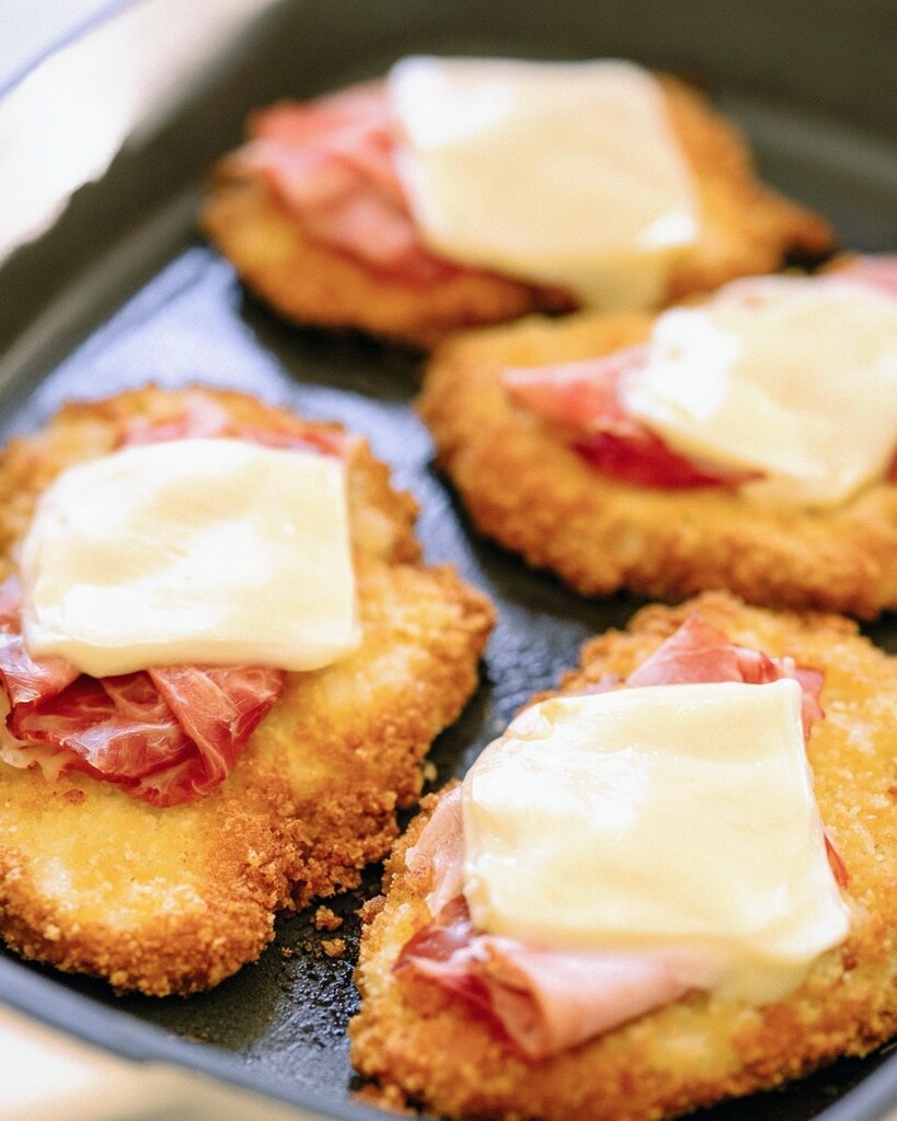 Golden chicken cutlets topped with folded sliced ham and white cheese resting in a black skillet