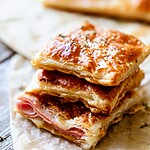 Several squares of baked ham and cheese puff pastry stacked on a piece of parchment paper.