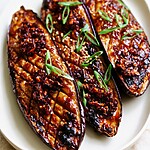 Tender Miso Glazed Eggplant with a Chili Crisp Finish Miso glazed eggplant halves topped with chili crisp, scallions, and sesame seeds on a white plate