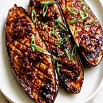 Tender Miso Glazed Eggplant with a Chili Crisp Finish Miso glazed eggplant halves topped with chili crisp, scallions, and sesame seeds on a white plate