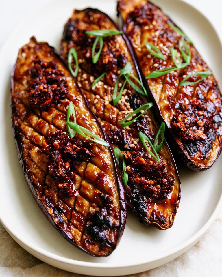 Tender Miso Glazed Eggplant with a Chili Crisp Finish Miso glazed eggplant halves topped with chili crisp, scallions, and sesame seeds on a white plate