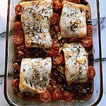 Fully baked white fish in a glass baking dish, topped with fresh dill and lemon zest, surrounded by soft tomatoes and olives