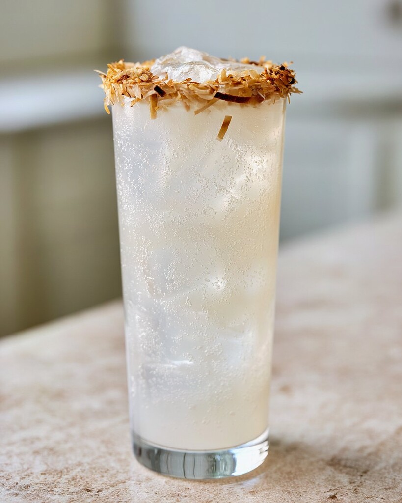 A tall glass of coconut lime vodka crush with a toasted coconut rim, surrounded by ice and a lime wedge