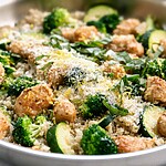 A metal skillet filled with cooked chicken pieces, fluffy quinoa, bright green broccoli florets, and zucchini, garnished with Parmesan and fresh basil.