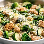 One-Pan Parmesan-Lemon Chicken and Quinoa A metal skillet filled with cooked chicken pieces, fluffy quinoa, bright green broccoli florets, and zucchini, garnished with Parmesan and fresh basil.