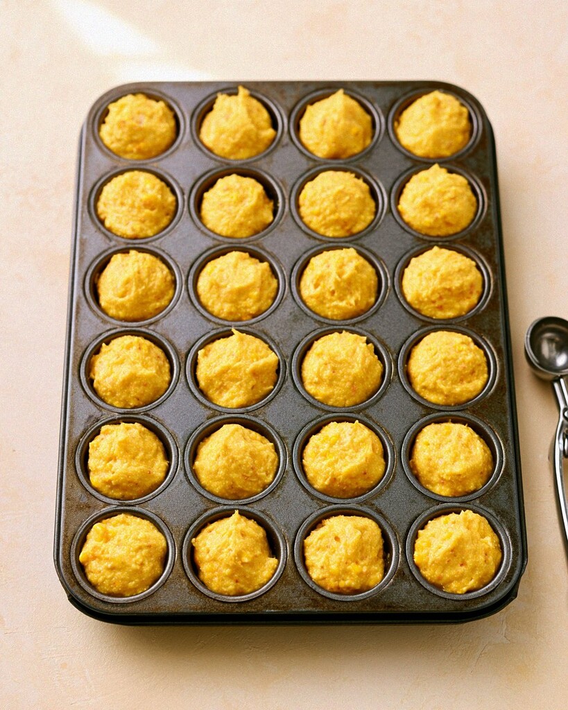 Thick yellow cornbread batter distributed evenly into the cavities of a metal mini muffin pan