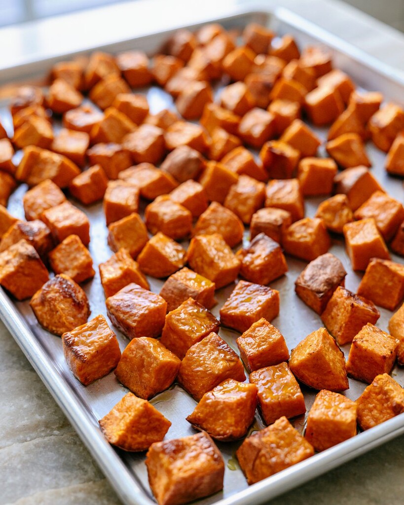 Glossy orange diced sweet potatoes roasting on a metal baking sheet with lightly browned edges