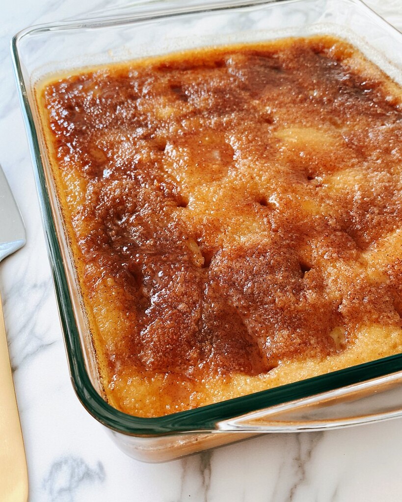 Wooden spoon handle poking holes into a golden sheet cake while a dark brown cinnamon syrup is poured over the surface