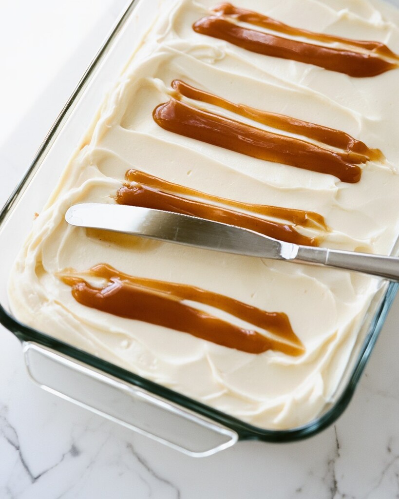 Brown cinnamon filling drizzled in straight lines over thick white cream cheese frosting inside a glass pan