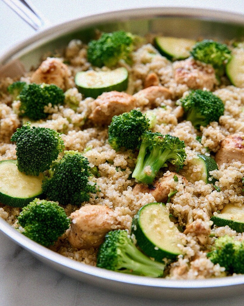 Broccoli florets, zucchini slices, and browned chicken pieces layered over partially cooked quinoa.