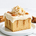 A square slice of cinnamon roll poke cake on a white plate topped with a cream rosette and dusted with cinnamon