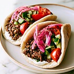 A warm pita pocket overflowing with spiced ground beef, diced cucumbers, tomatoes, and bright pink sumac onions on a ceramic plate