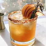 A rocks glass holding an amber cocktail and ice, edged with a red chili rim and garnished with apple slices and rosemary
