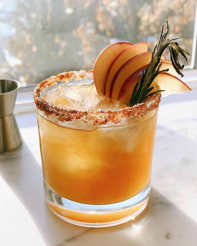 A rocks glass holding an amber cocktail and ice, edged with a red chili rim and garnished with apple slices and rosemary