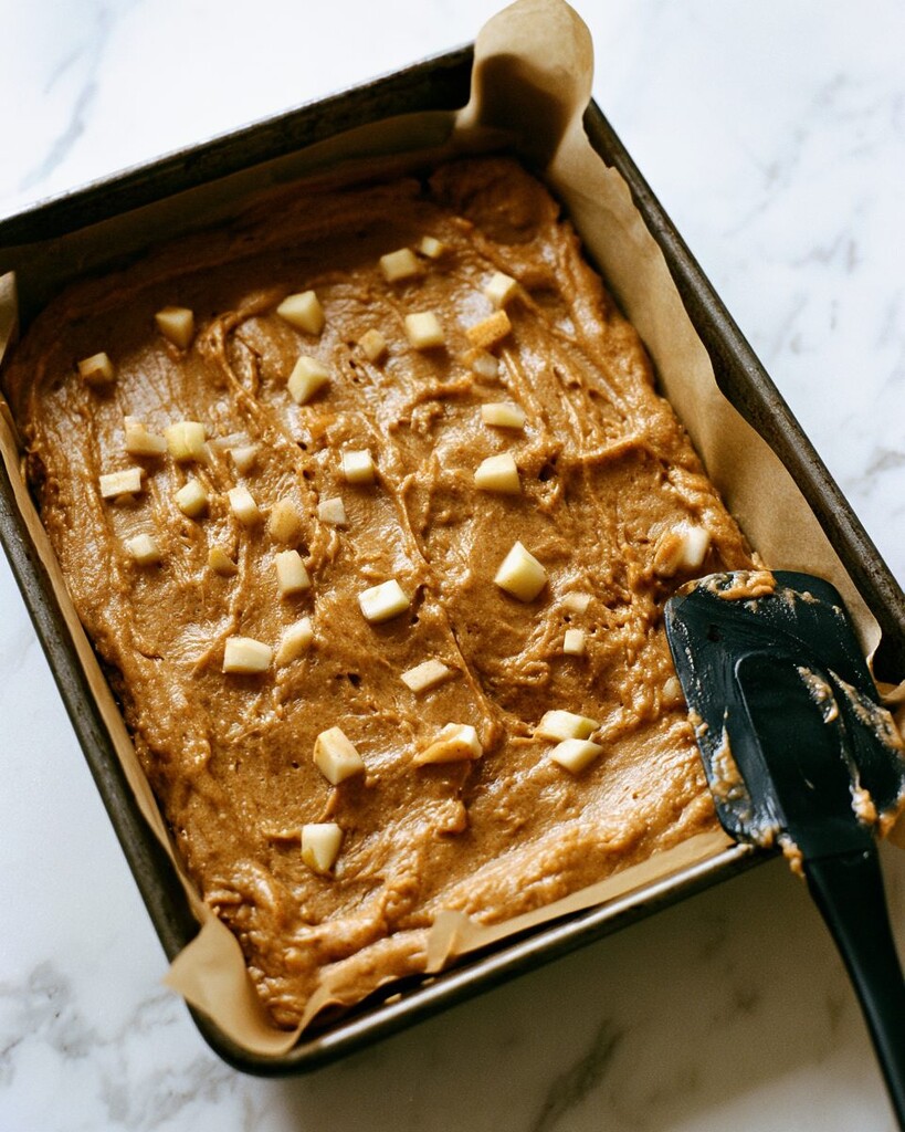 Unbaked blondie batter spread in a parchment-lined pan with diced apples scattered across the surface