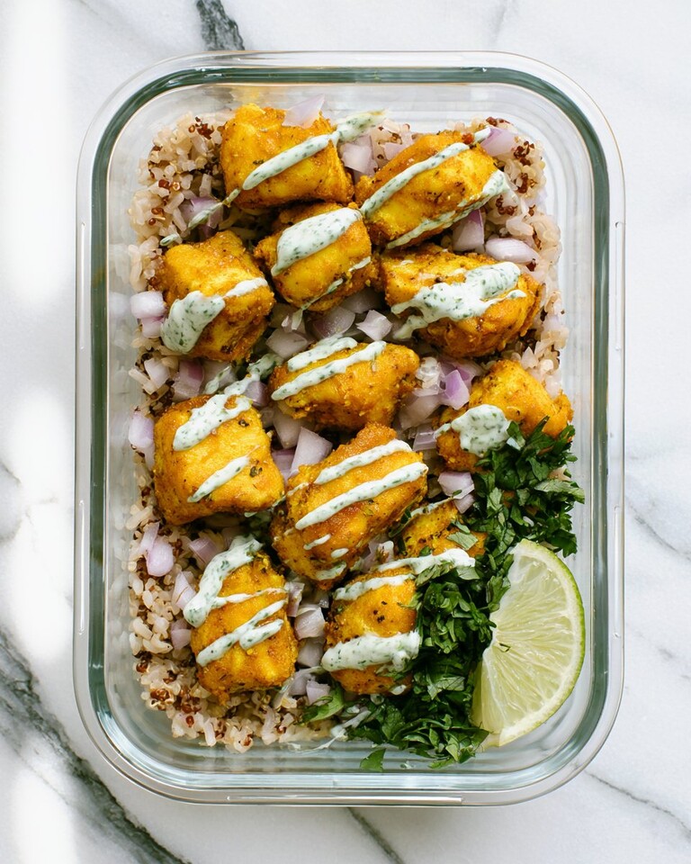 20-Minute Curried Chicken Meal-Prep Bowls Glass bowl holding golden curried chicken over rice with cilantro-lime yogurt sauce and red onions
