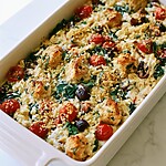 Baked Greek chicken and orzo casserole topped with golden panko, fresh basil, and roasted cherry tomatoes in a white dish
