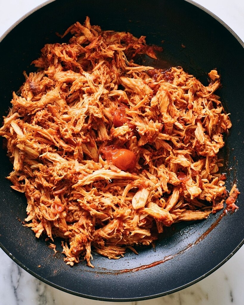 Shredded chicken being tossed with thick red salsa inside a skillet