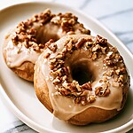 Close up of a glazed baked apple cider doughnut with creamy maple icing and chopped pecans on a light plate