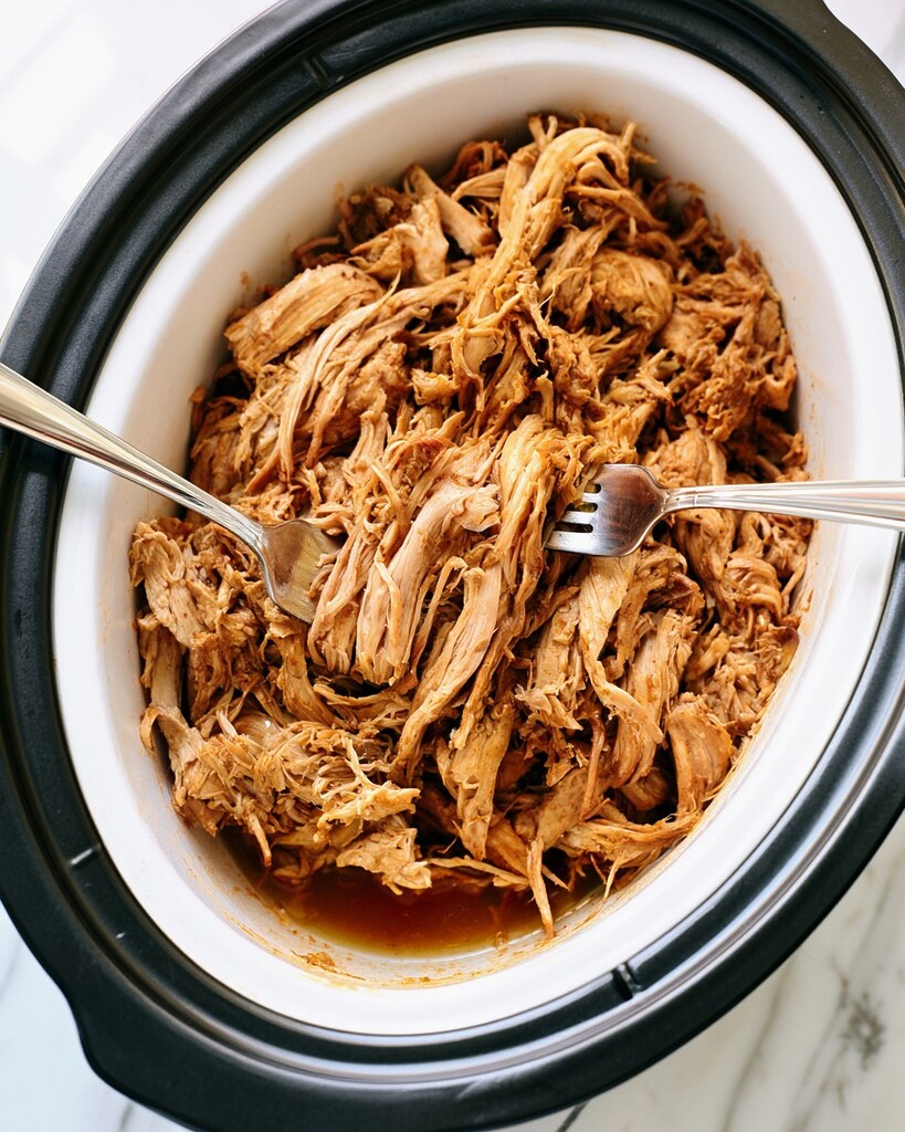 Shredding cooked chicken breasts into tender strips inside a slow cooker using two metal forks