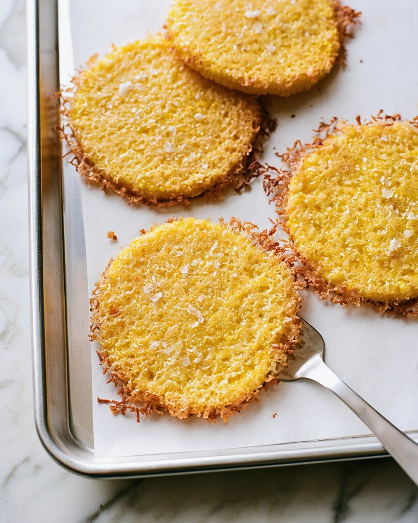 Lifting a baked polenta bite off the parchment paper with a metal spatula to show the thin crisp cheese crust