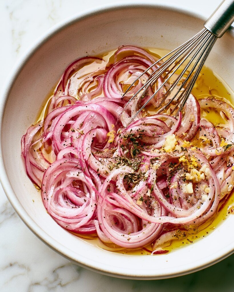 Thinly sliced red onions soaking in lemon juice inside a metal mixing bowl