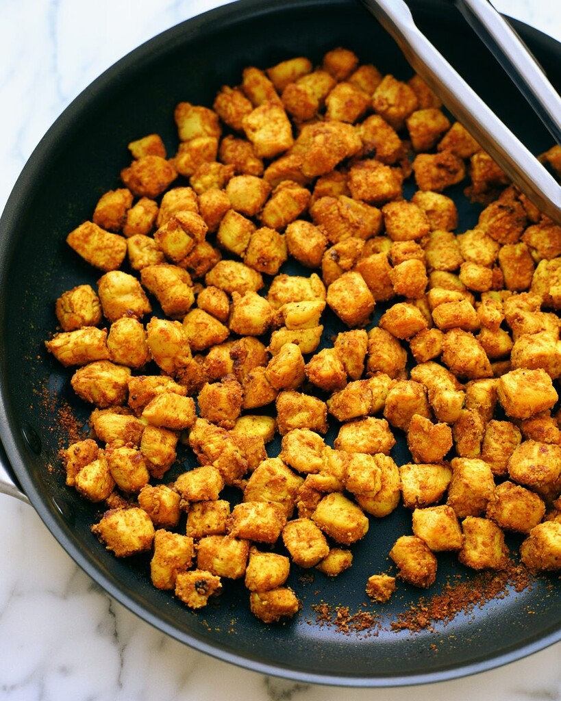 Golden curried chicken pieces searing in a black skillet held by metal tongs