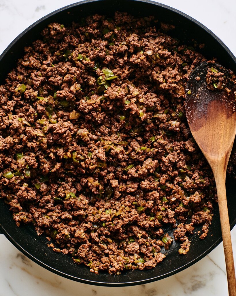 Ground beef and diced onions browning in a black skillet, broken apart with a wooden spoon
