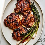 Plated huli huli chicken thighs on a light background with charred scallions and sesame seeds