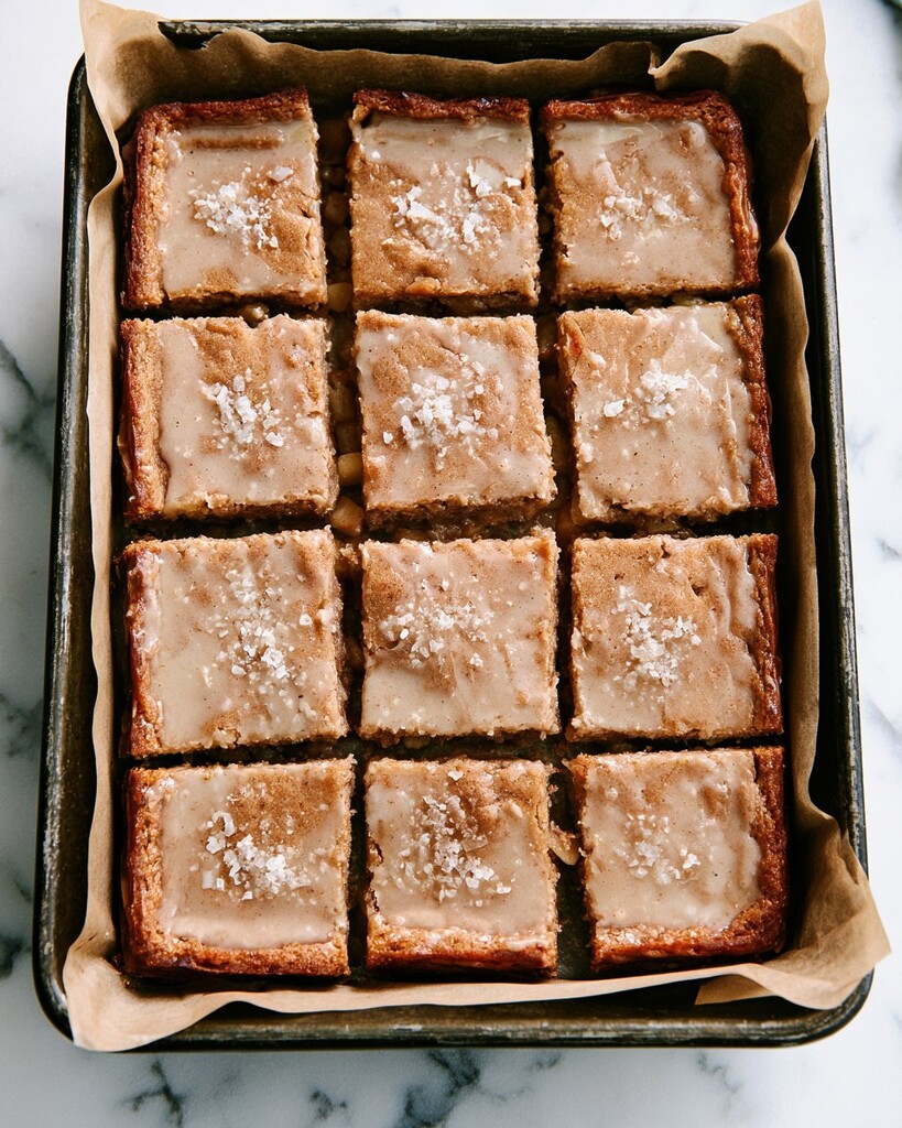 Sliced squares of glazed apple blondies inside a lined baking pan finished with flaky sea salt