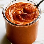 Thick, smooth reddish-brown pumpkin butter being transferred into a glass jar with a serving spoon