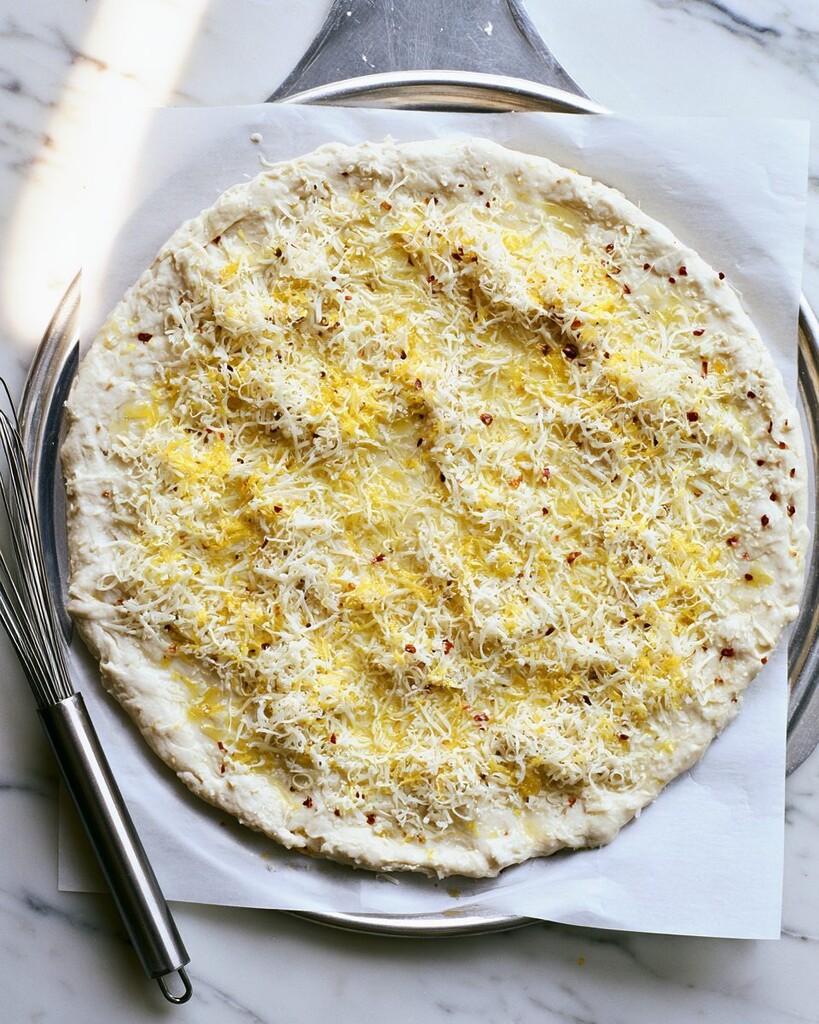 Unbaked pizza dough topped with shredded cheese, lemon zest, and red pepper flakes on a metal pan