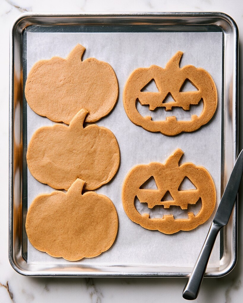Pumpkin shaped cookie dough cutouts on a parchment lined baking sheet, half plain and half with carved faces