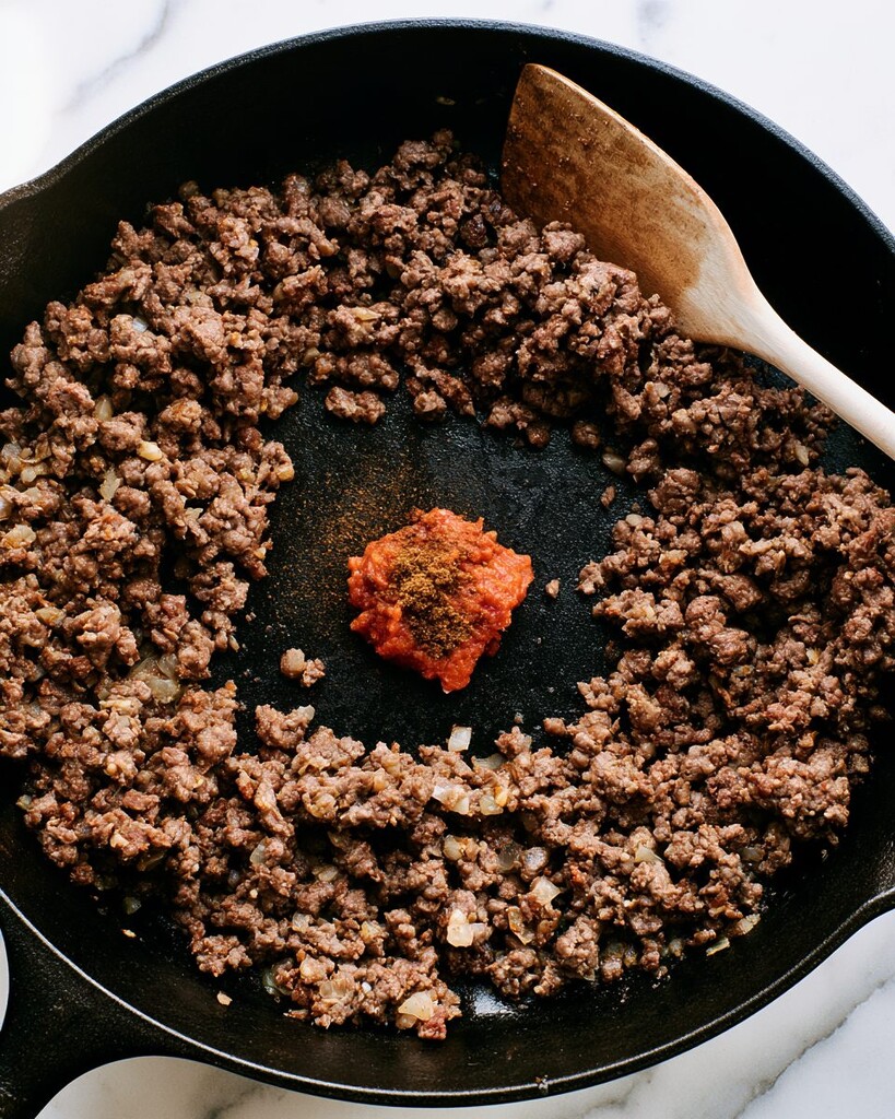 Ground beef pushed to the edges of a cast iron skillet with tomato paste and dark spices toasting in the center