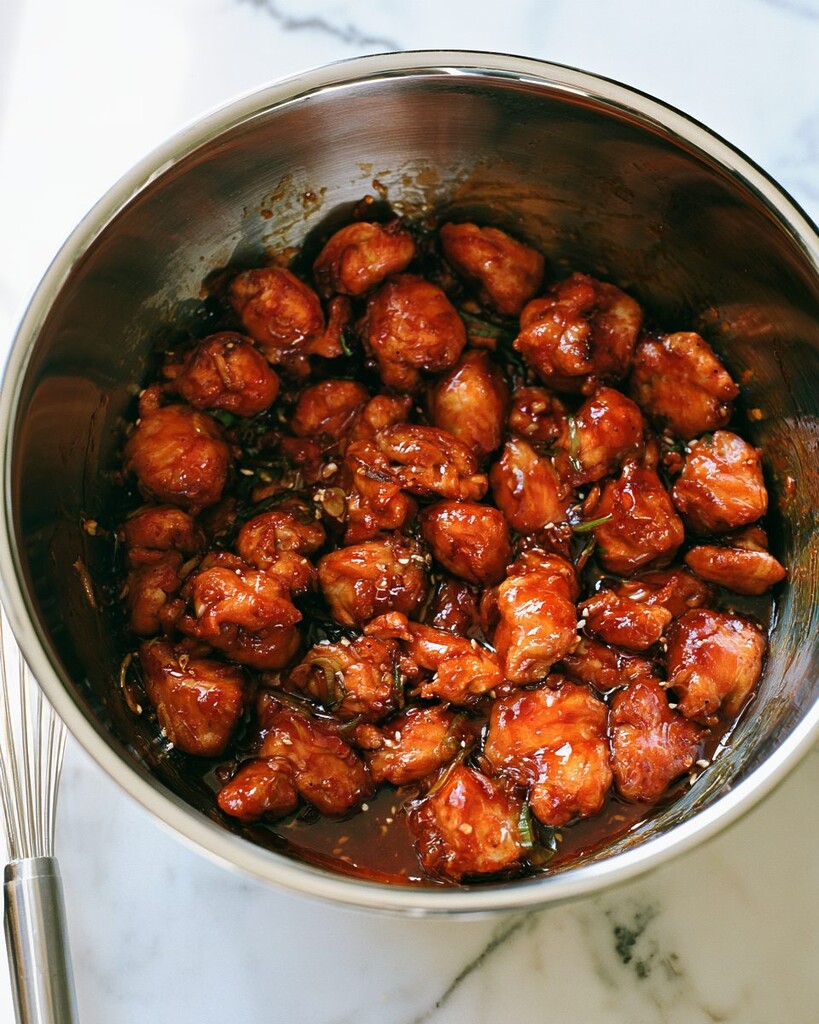 Stainless steel mixing bowl filled with hot chicken pieces heavily coated in a glossy honey and sriracha sauce