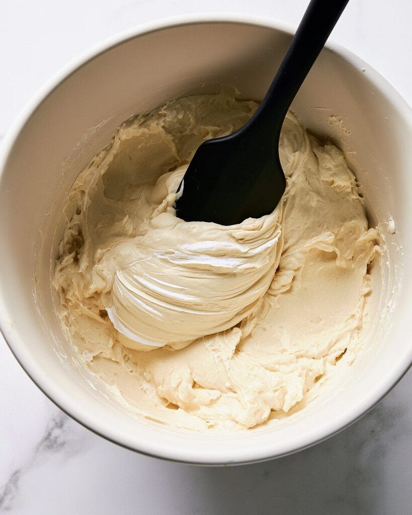 Thick cheesecake mousse being folded with a silicone spatula in a white bowl
