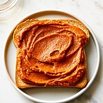 Hands-Off Oven-Roasted Vanilla Bean Pumpkin Butter Dark reddish-brown vanilla bean pumpkin butter spread thickly over a slice of toasted bread resting on a light-colored plate