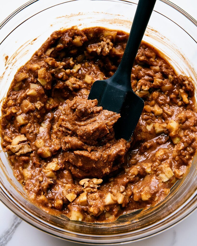 Folding diced apples and chopped pecans into dark blondie batter in a glass bowl with a silicone spatula