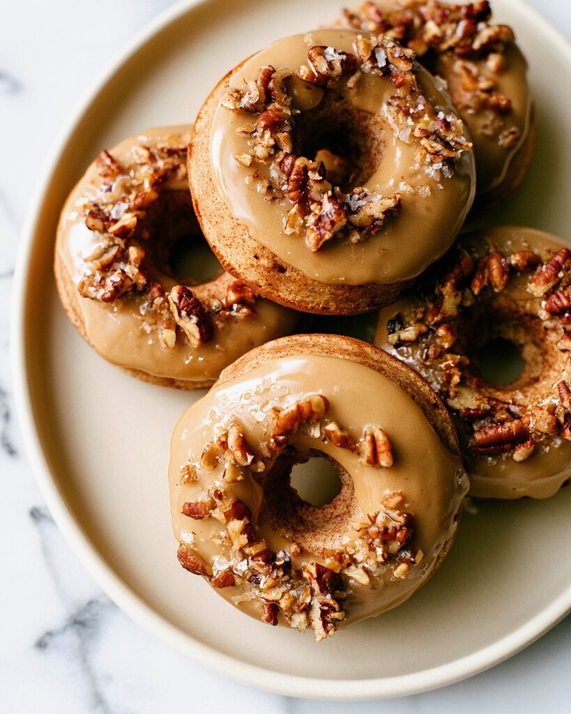 Stacked baked apple cider doughnuts on a light plate with chopped pecans and maple glaze