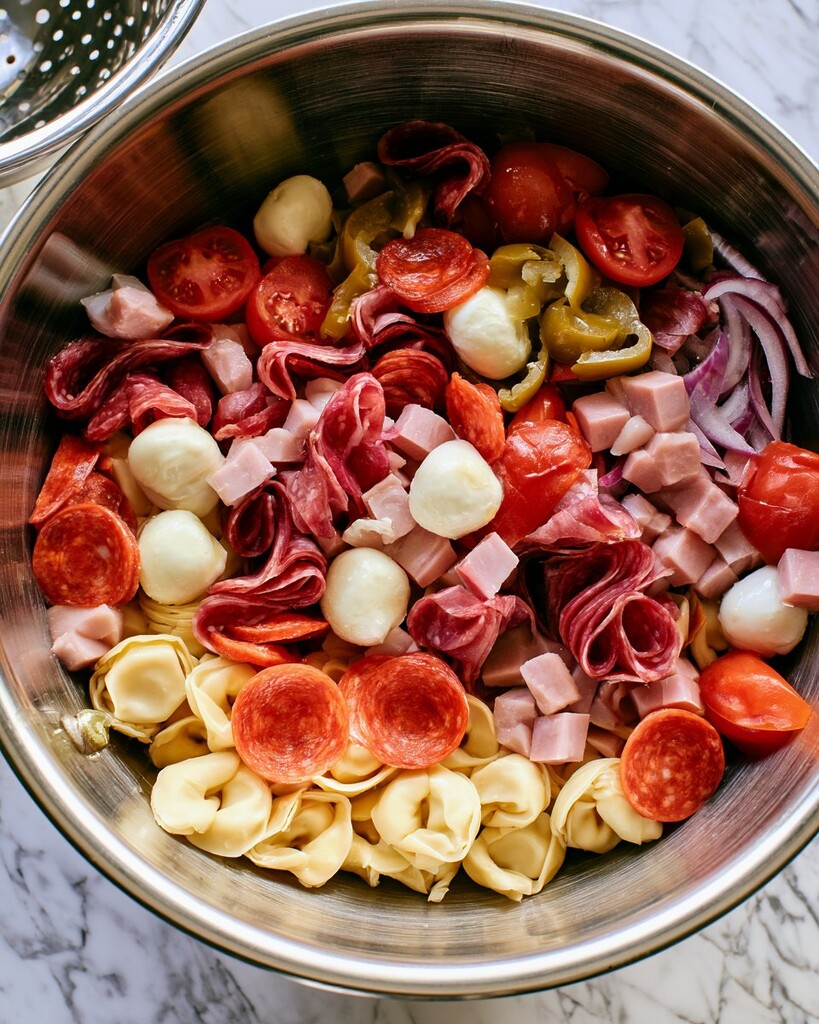 Cooked tortellini, sliced salami, pepperoni, ham, mozzarella, tomatoes, and red onion layered in a metal bowl