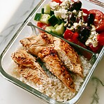 Greek Chicken Rice Bowls with 2-in-1 Dressing Greek chicken rice bowl in a glass container with cherry tomatoes, cucumber, and feta salad