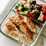 Greek Chicken Rice Bowls with 2-in-1 Dressing Greek chicken rice bowl in a glass container with cherry tomatoes, cucumber, and feta salad
