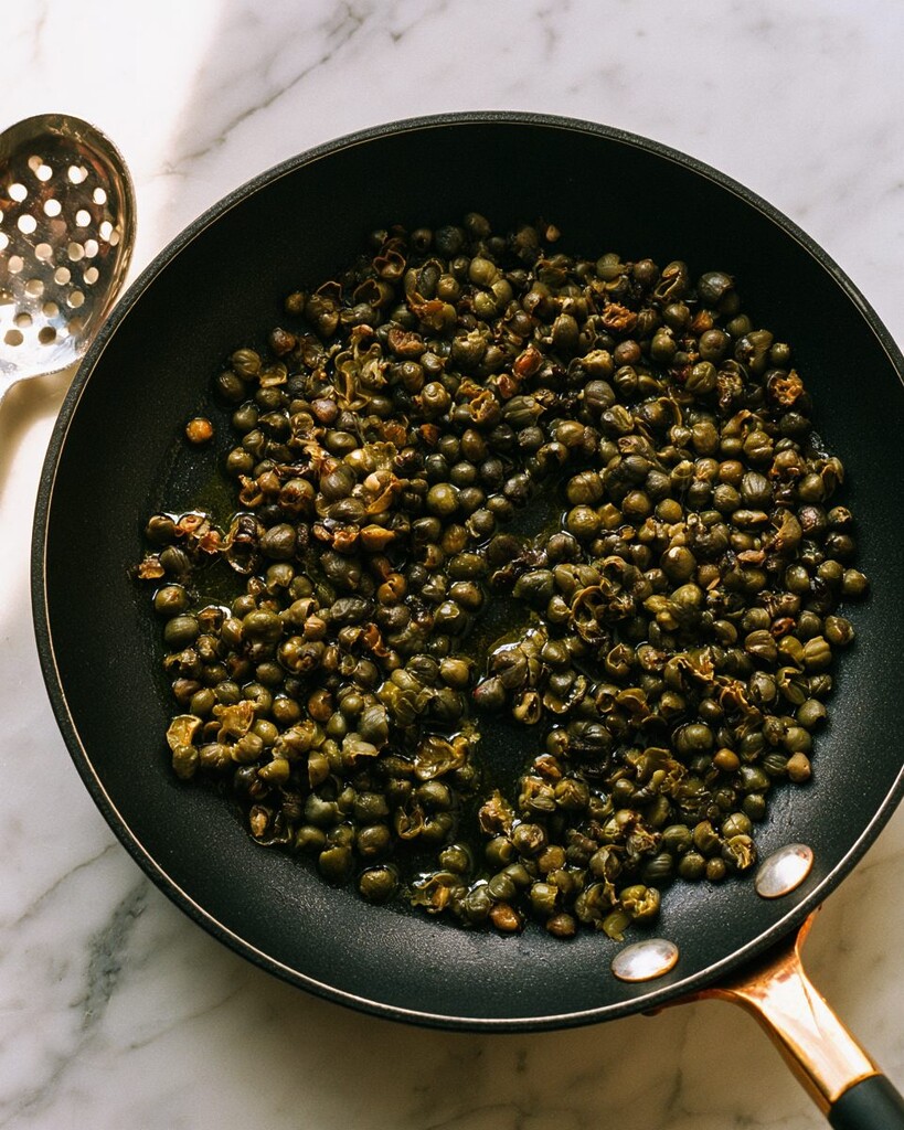 Capers frying in a black skillet until they burst open and turn dark and crispy