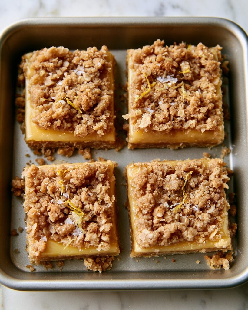 Four brown butter lemon crumb bars arranged on a small tray, topped with crisp oat topping and fresh zest