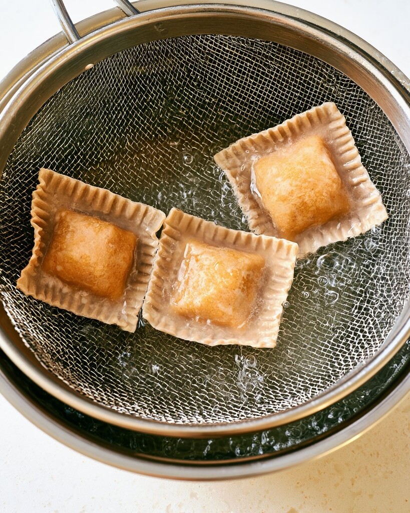 Translucent wonton ravioli being lifted from boiling water with a metal mesh spider