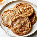 Browned butter salted honey cookies arranged on a light plate with golden crinkled surfaces and scattered flaky salt