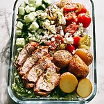 Glass meal prep bowl with roasted Greek chicken, potatoes, chopped romaine, and feta cucumber salad
