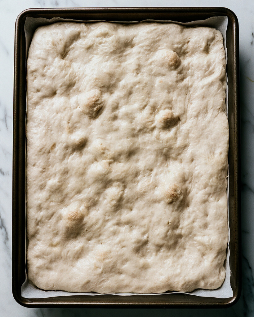 Cinnamon roll focaccia dough filling a parchment-lined pan showing large air bubbles on the surface